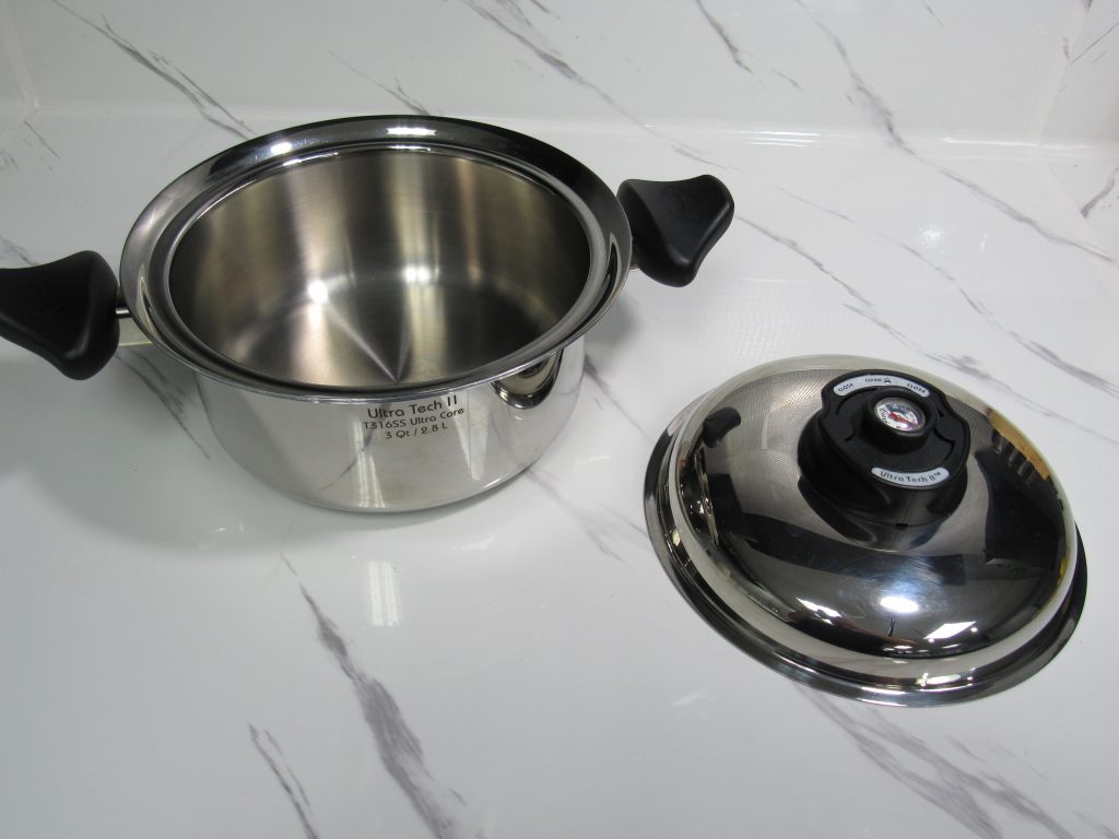 The Food You Eat 2 Titanium steel 3-quart Ultra Tech II saucepan with black handles, shown with matching lid on a white marble countertop.