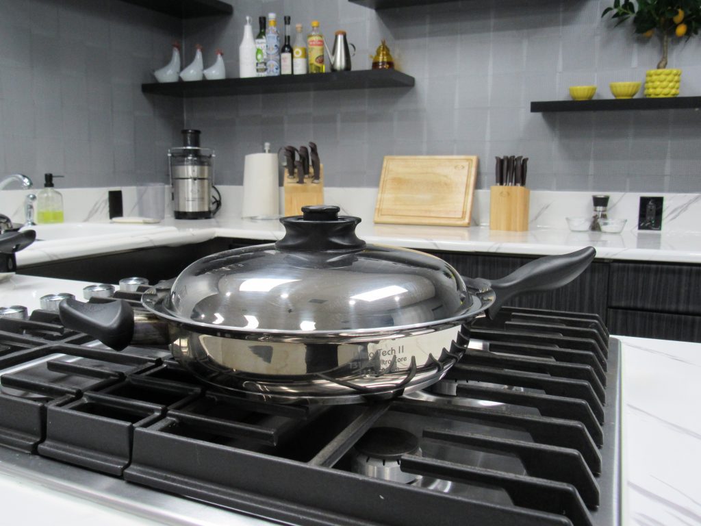 The Food You Eat 1 Titanium steel skillet with lid on a gas stove in a modern kitchen with gray tile backsplash, marble countertops, and cooking utensils.