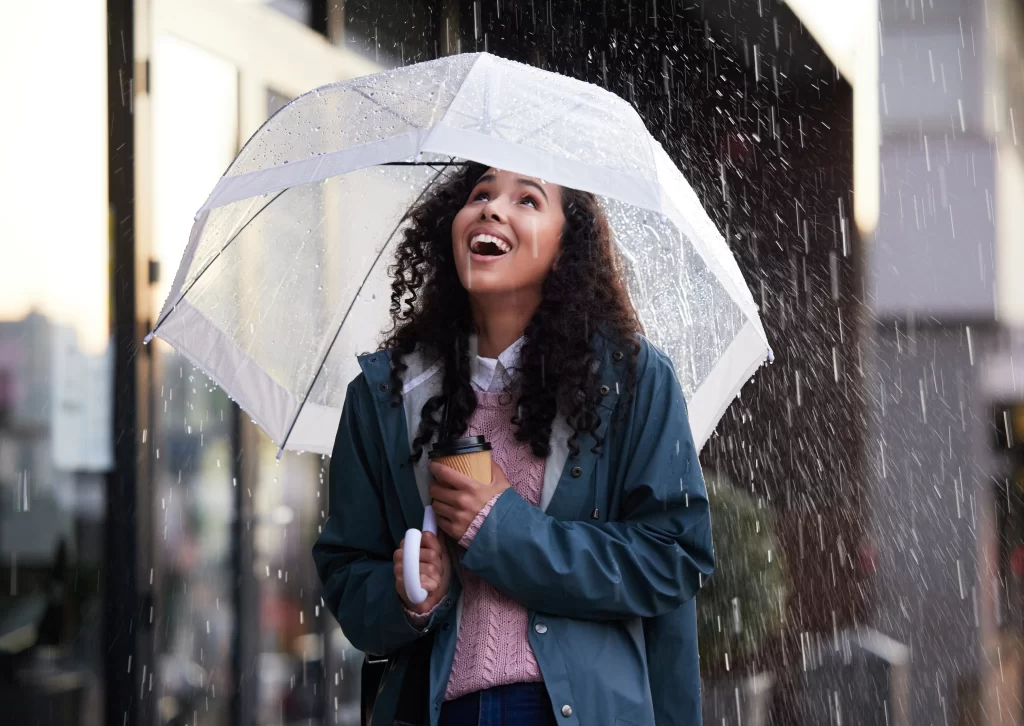 A smiling woman holding a coffee cup under a clear umbrella during a rainstorm, highlighting how seasonal weather affects a home water filter system.