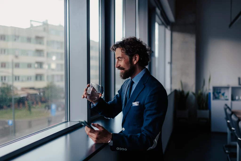 A businessman in a suit looking out an office window while drinking a glass of clean water filtered by a whole house water purification system.