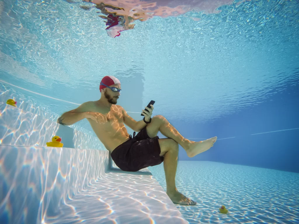 Coming Home: The One Vacation Souvenir You Can't Pack 2 A man wearing a swim cap and goggles sits relaxed on the steps underwater in a crystal-clear swimming pool, looking at his smartphone next to yellow rubber ducks.