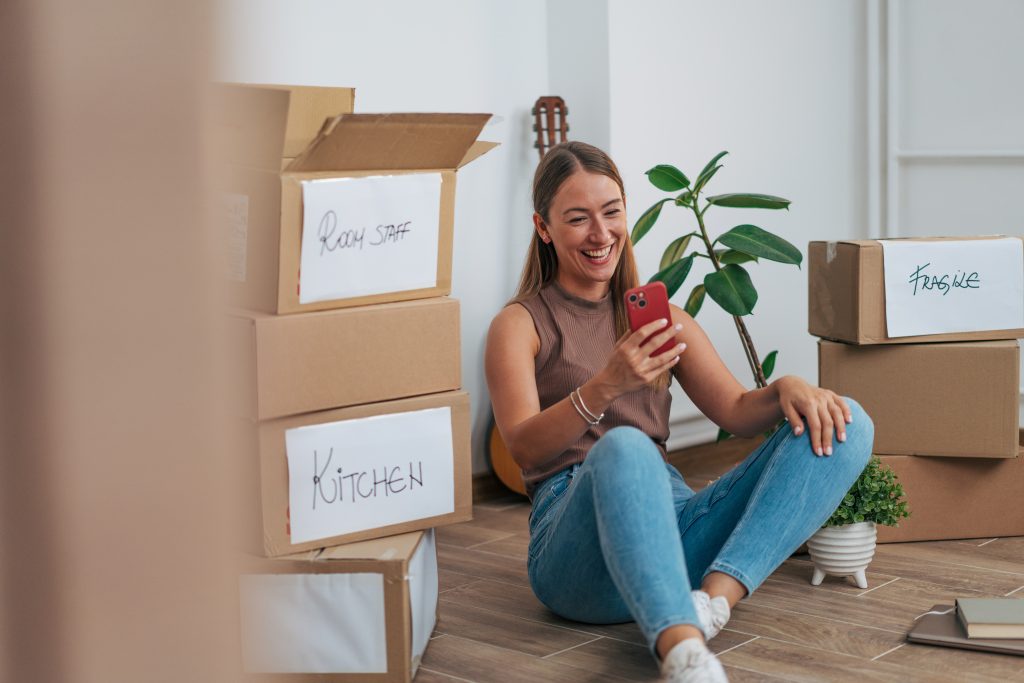 Starting Fresh: Building a Healthy Home from Day One 2 Young woman smiling on her phone surrounded by labeled moving boxes in a new home